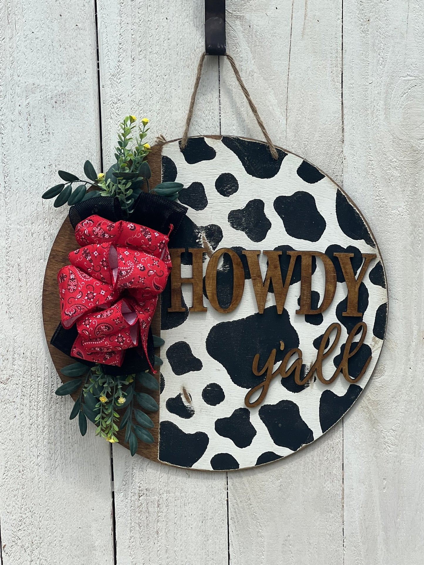 Cow Print Howdy Y’all Wood Door Hanger in black, red, and white with rustic charm hanging on a white fence.