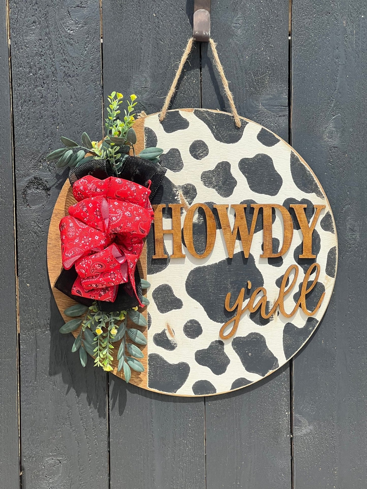 Cow Print Howdy Y’all Wood Door Hanger in black, red, and white with rustic charm hanging on a black fence.