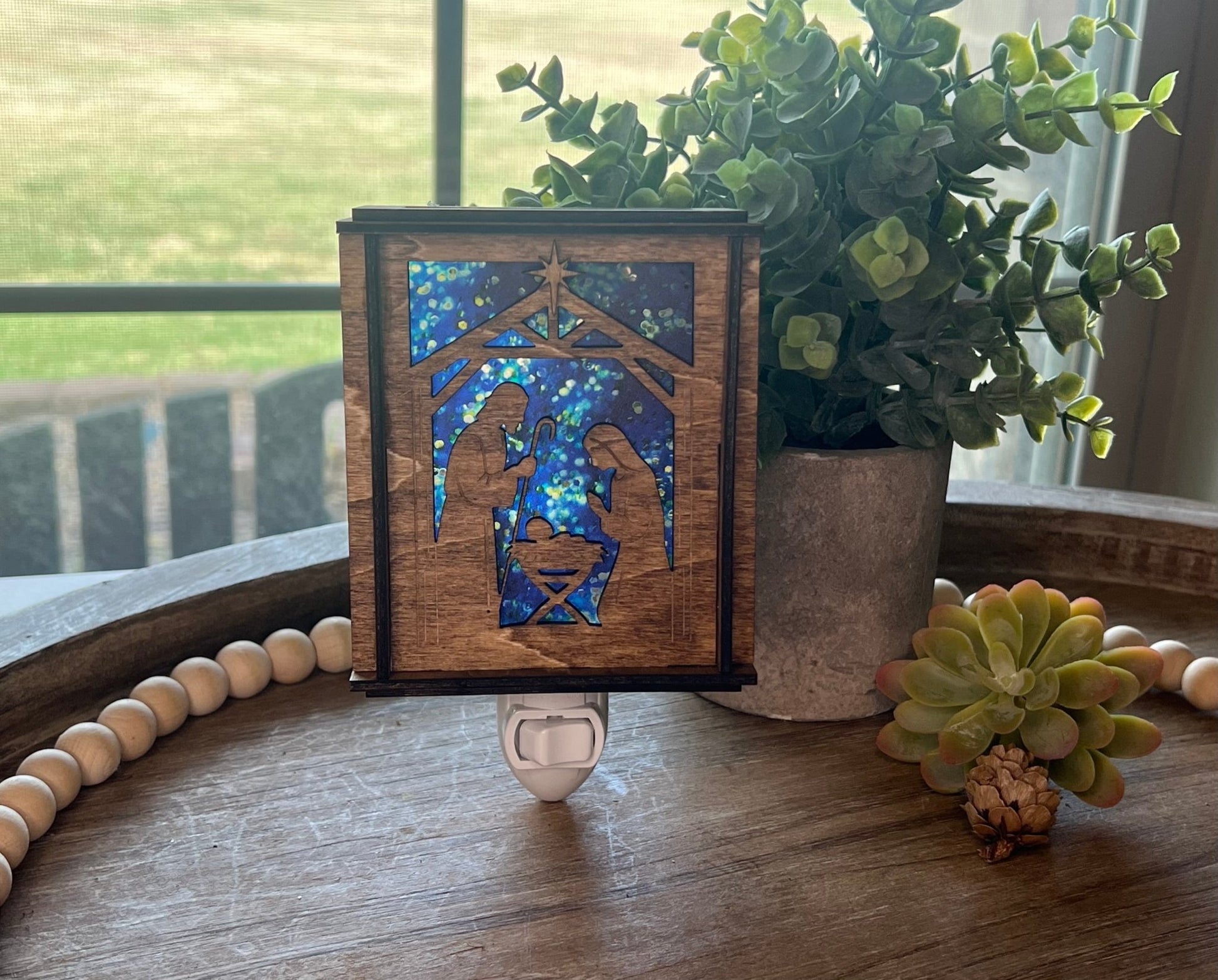 A Christmas Nativity Lantern Nightlight in golden oak with wood and acrylic materials on a try with natural light shining through.