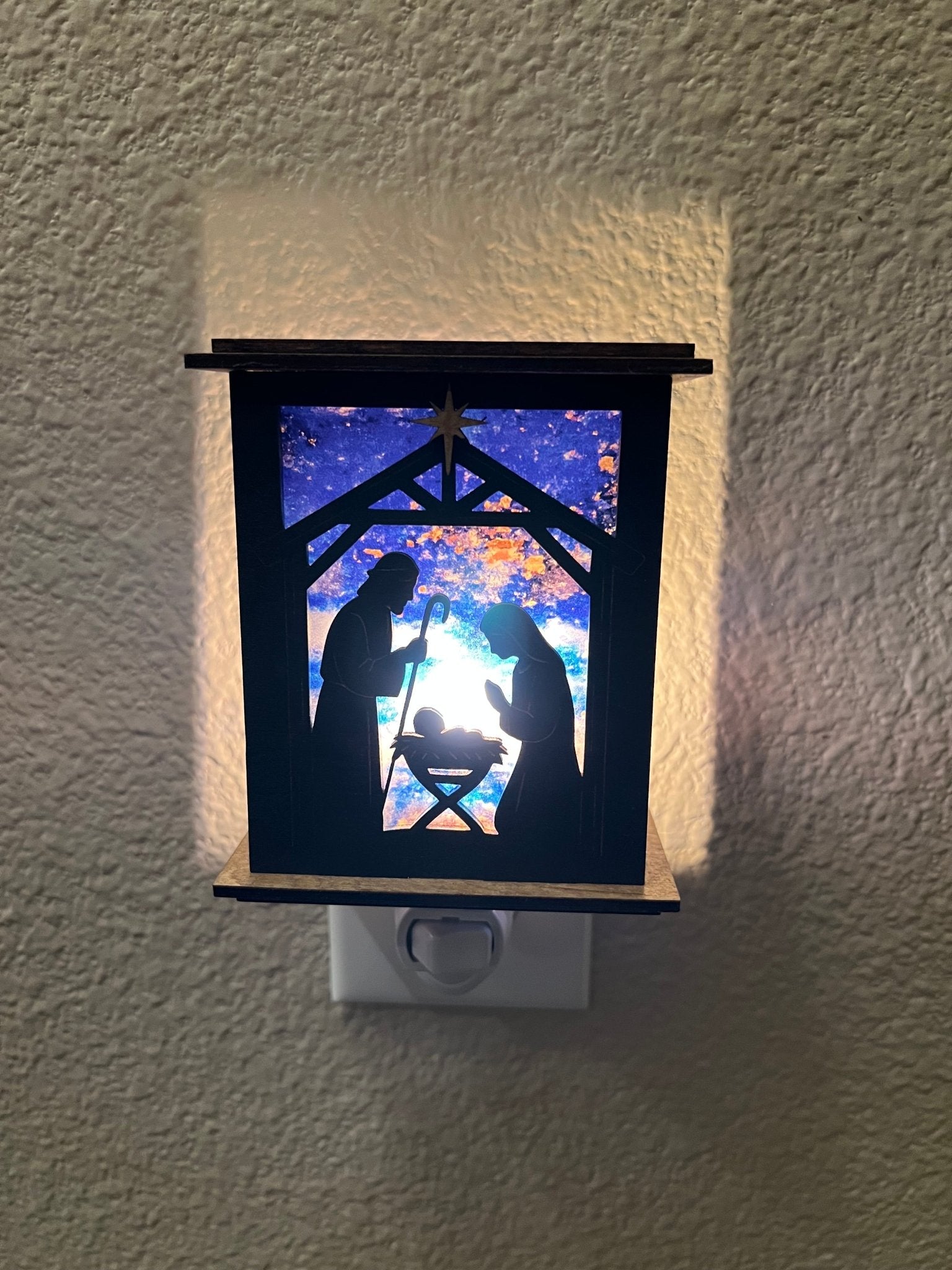 Rustic nativity nightlight lantern made of black wood and golden oak stained acrylic plugged in with front view and lit.