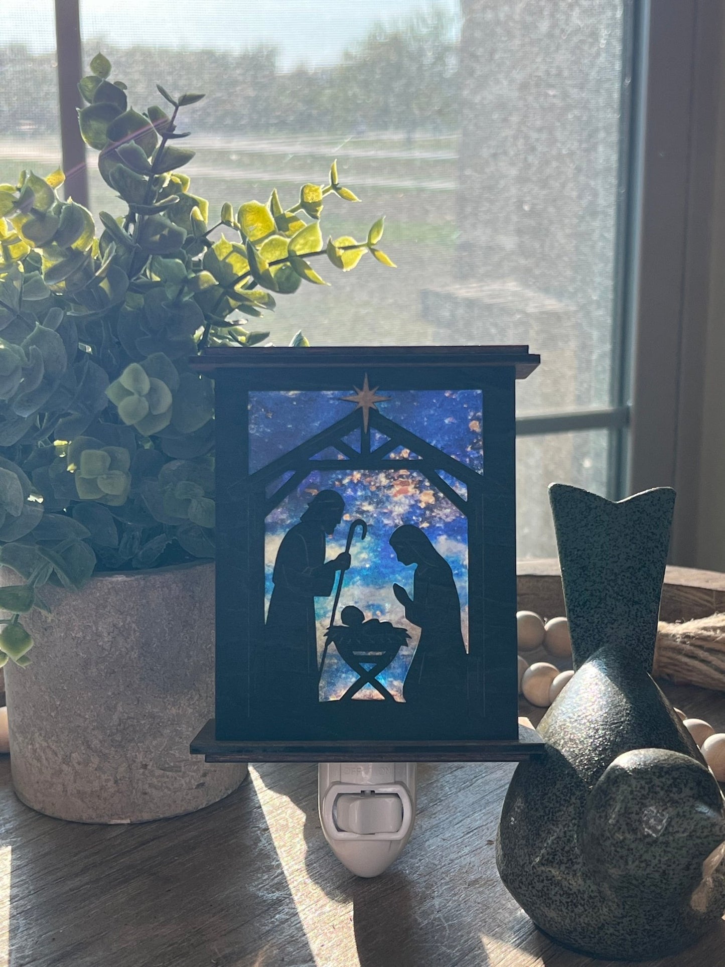 Rustic nativity nightlight lantern made of black wood and golden oak stained acrylic standingup  with front view and sunlit.